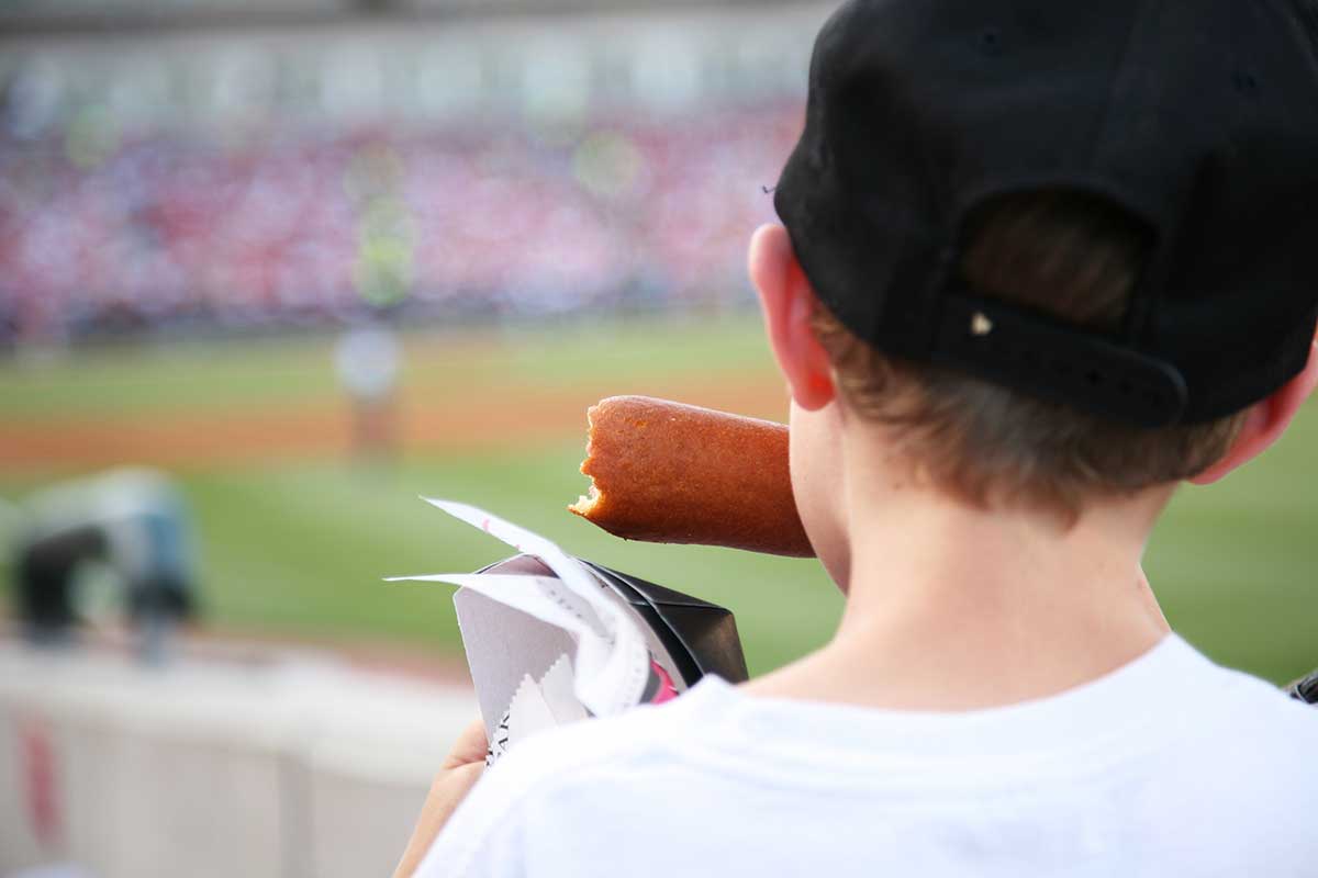 little boy at a baseball game eating a corndog